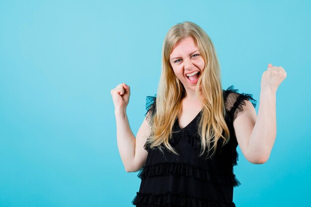 Screaming happy girl is raising up her fists on blue background