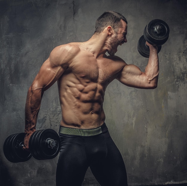 Screaming, shirtless muscular male working out with dumbbells on grey background.