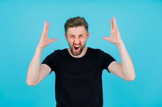 Screaming young man is raising up his hands on blue background