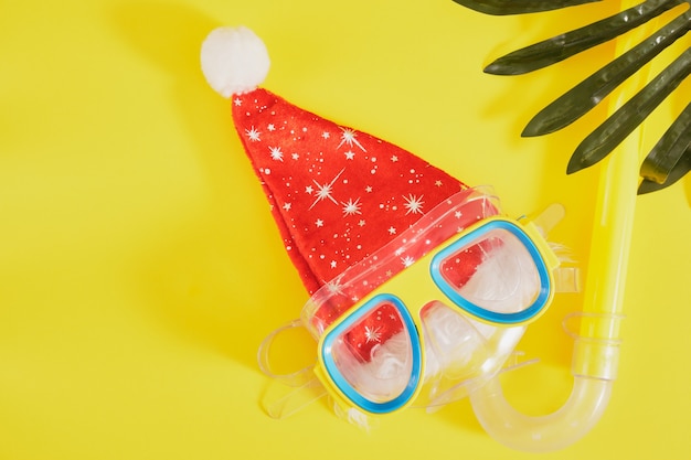 Набор для подводного плавания, шляпа санта-клауса и пальмовый лист на желтом фоне, рождественские каникулы на пляже в теплой стране