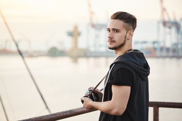 Sea attracts this photographer. Outdoor portrait of attractive young guy standing in harbour, enjoying looking at sea while holding camera, searching for good location to take photo, looking aside