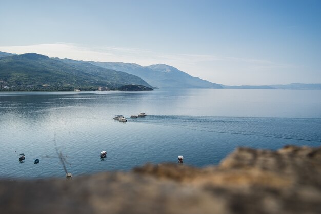 Sea with boats on it surrounded by hills covered in greenery under sunlight