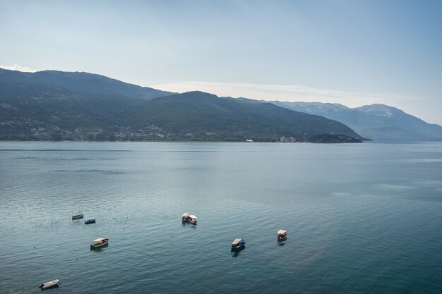 Sea with boats on it surrounded by hills covered in greenery under sunlight