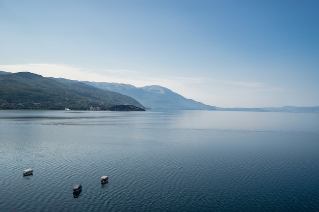 Sea with boats on it surrounded by hills covered in greenery under sunlight