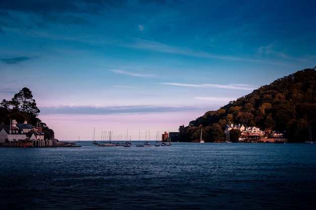 A sea with ships and  buildings on the coast surrounded by high green mountains in Dartmouth, UK
