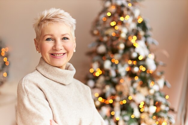 季節、冬、休日、お祝いのコンセプト。自宅で飾られた松の木でポーズをとって、クリスマスの準備を楽しんでいる短い髪と広い輝く笑顔を持つ陽気な中年の女性の写真