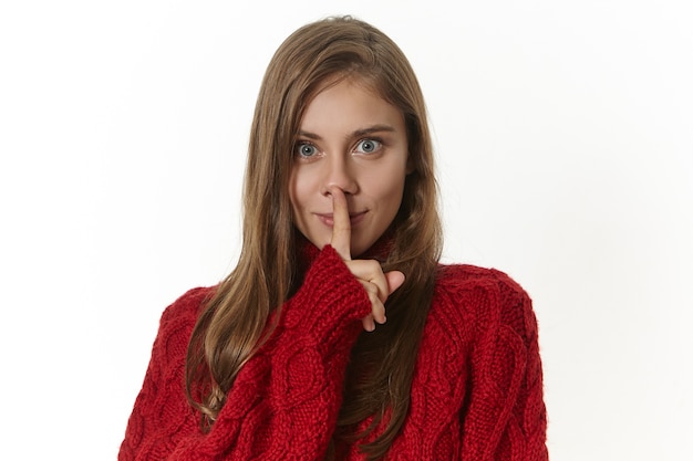 Secrecy, conspiracy and confidential information concept. Isolated image of attractive mysterious young lady in sweater making shh geture with fore finger at lips, asking to keep her secret