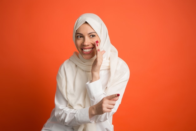 秘密のゴシップコンセプト。ヒジャーブの幸せなアラブの女性。赤いスタジオの背景でポーズをとって、笑顔の女の子の肖像画。若い感情的な女性。人間の感情、表情のコンセプト。正面図。