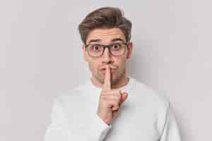 Free photo secret young unshaven man makes silence gesture preses indxx finger over lips says shh wears spectacles and casual jumper demands to be quiet isolated over white background. body language concept