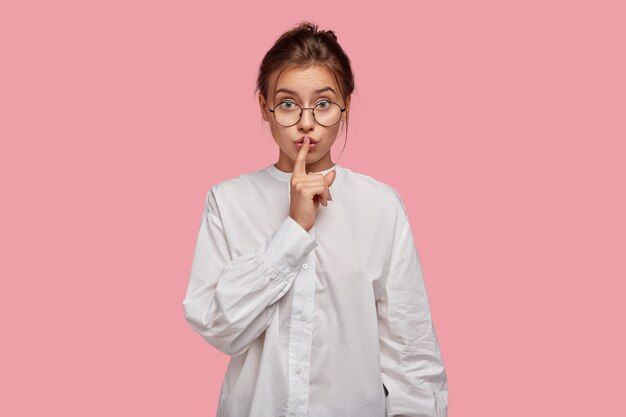 Secretive young woman with glasses posing against the pink wall