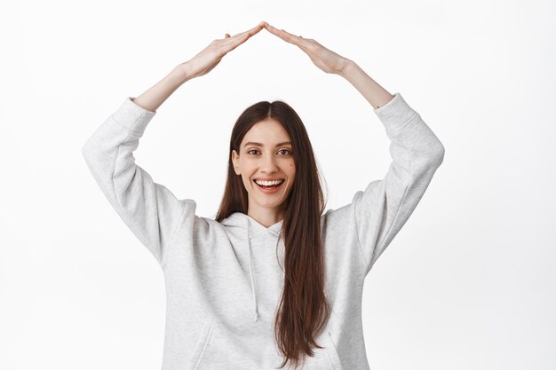 Secure your home. Smiling young woman showing roof, rooftop gesture with hands above head, protect herself, looking happy and determined at camera, white background