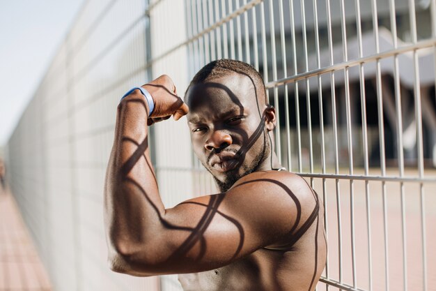 Соблазнительный афроамериканец с мускулами позирует с обнаженным сундуком перед забором