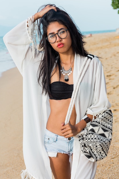 Seductive Asian woman in black top and jeans shorts , beach cover up with boho bag posing on tropical beach. Bohemian woman enjoying weekends. Perfect body.Jewelry , bracelet and necklace.