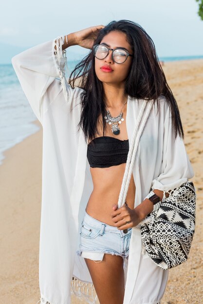 Соблазнительная азиатская женщина в черном топе и джинсовых шортах, пляж прикрывает сумкой бохо, позируя на тропическом пляже. Богемная женщина, наслаждающаяся выходными. Идеальное тело. Украшения, браслет и колье.