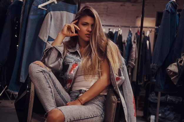 Free photo seductive girl wearing a distressed jeans and denim jacket looking at a camera while sitting on a chair in the fitting room of a clothing store.