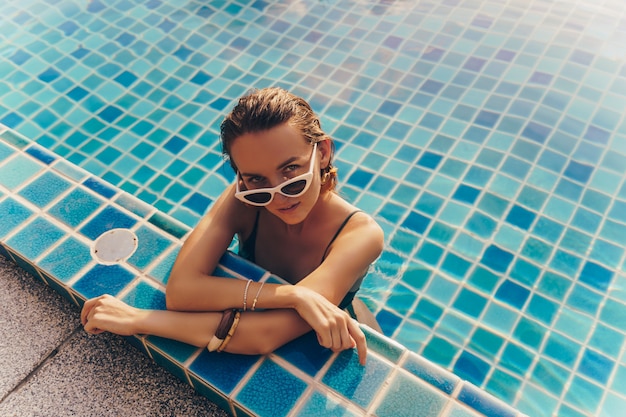 Seductive graceful woman in stylish yellow earrings with perfect body posing in the pool during holidays on luxury resort