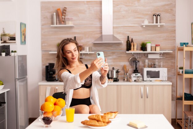 自宅のキッチンでスマートフォンを使用して自分撮りをしているセクシーなランジェリーの魅惑的な女性。朝に魅力的な下着を着てスマートフォンを使用して入れ墨を持つ魅力的な女性。