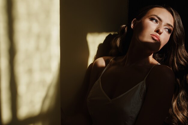 Seductive young bride in a silk dress sits on the windowsill in the early morning