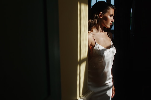 Seductive young bride in a silk dress sits on the windowsill in the early morning