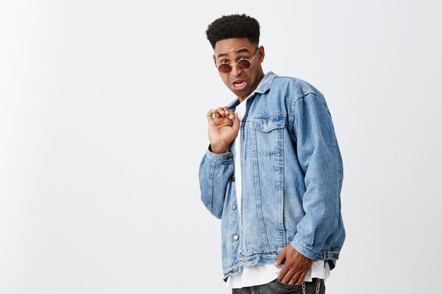 Seeing ex girlfriend with ugly guy. Portrait of young attractive black-skinned man with afro haircut in white shirt, denim jacket and tan glasses looking in camera with disgust expression.