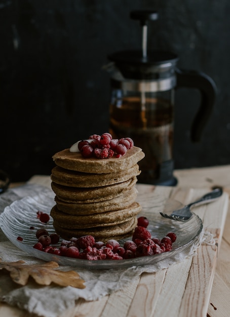 Selective closeup shot of a stack of pancakes with red berries on a plate near a tea pitcher