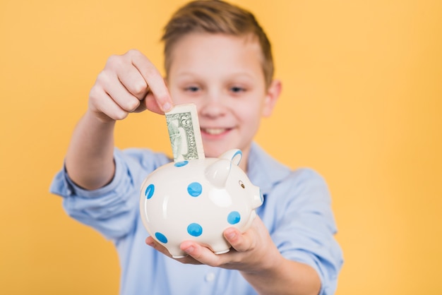 Selective focus of a boy inserting currency note in the polka dot ceramic piggy bank against yellow backdrop