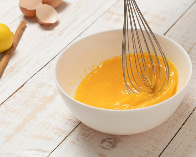 Selective focus closeup of a bowl of egg and an egg beater on a white plank table