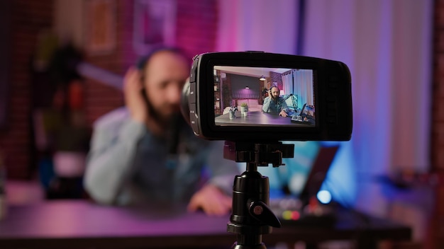 Selective focus on digital video camera screen recording male vlogger with headphones talking at microphone in home podcasting studio. Closeup of dslr filming influencer streaming live internet show.