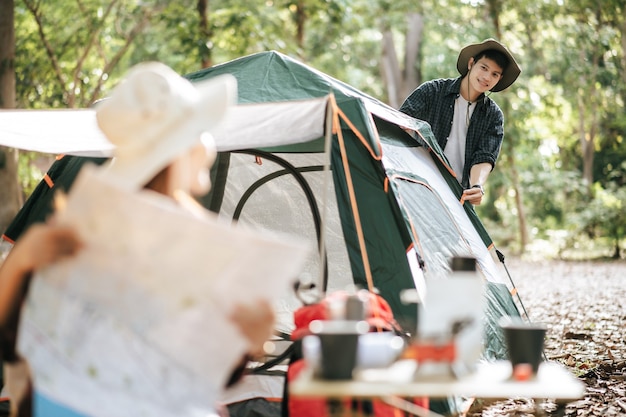セレクティブフォーカス、キャンプテントの前の椅子に座って紙の地図で方向を確認しているきれいな女性、彼女の後ろにテントを張っているハンサムなボーイフレンド、彼らは休暇で森でキャンプするのを喜んでいます