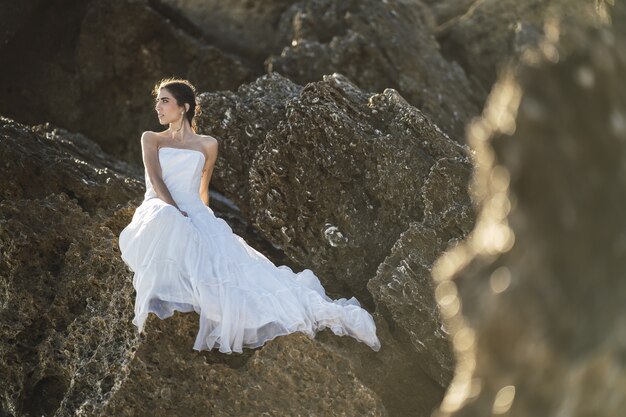 Selective focus shot of a brunette female in a white dress posing on the rocks