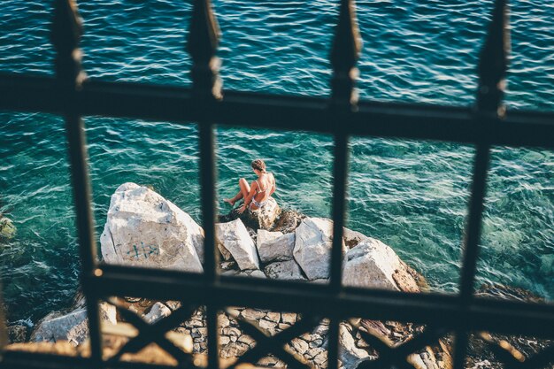 Foto gratuita colpo di messa a fuoco selettiva di un bikini femminile che si siede su una roccia dal corpo idrico