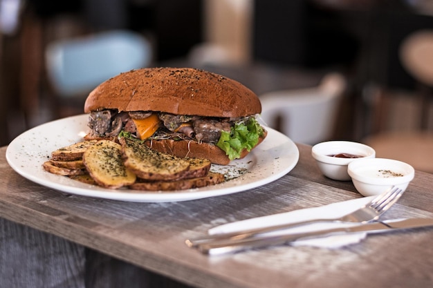 Selective focus shot of a tasty sandwich with potatoes on a wooden table with sauces and cutlery