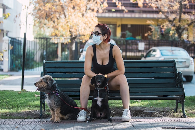Selective focus shot of a woman in a mask sitting on a bench with dogs