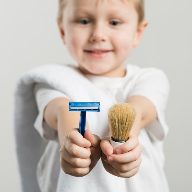 Selective focus of a smiling boy showing razor and shaving brush toward camera