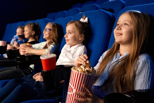 Selective focus of smiling little girl holding popcorn bucket, sitting with laughing friends in comfortable chairs in cinema. Children watching cartoon or movie, enjoying time