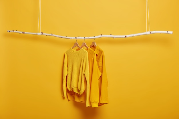 Selective focus. Three items of clothes on hangers. Long sleeved yellow jumpers on wooden rack near bright vivid wall.