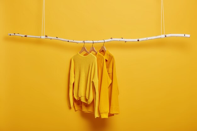Селективный фокус. Три предмета одежды на вешалках. Желтые перемычки с длинными рукавами на деревянной стойке возле яркой яркой стены.