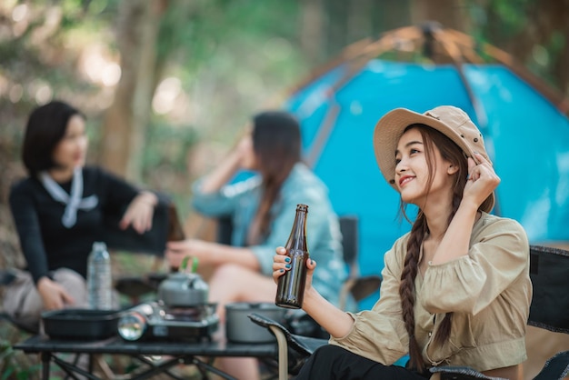 Избирательный фокус Молодая азиатская красивая женщина и ее подружки путешественники отдыхают в походных стульях в палатке. Они веселятся и пьют пиво во время кемпинга, разговаривая весело и счастливо вместе