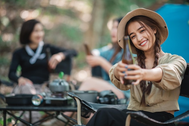 Selective focus young Asian pretty woman and her girlfriend sitting at front of tent use mobile phone take photo during camping in forest with happiness together