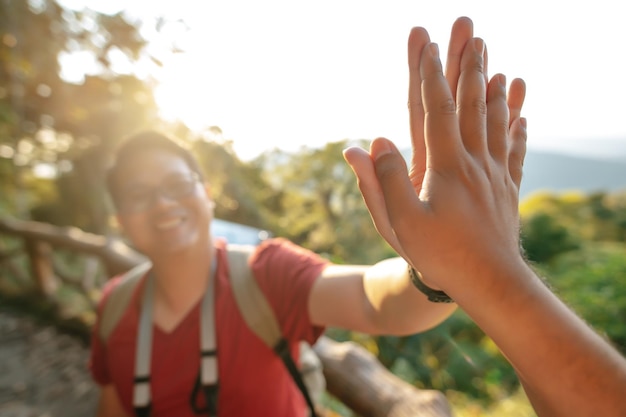 Free photo selective focus young backpacker man in glasses greeting giving high five clapping hands with someone in forest after finished on the rocky peak backlit the sunlight copy space