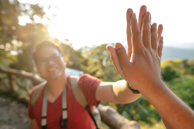 Selective focus Young backpacker man in glasses greeting giving high five clapping hands with someone in forest after finished on the rocky peak backlit the sunlight copy space