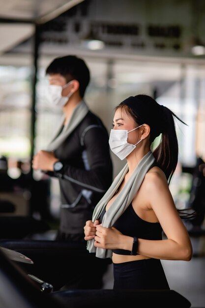 Free photo selective focus,  young sexy woman in mask wearing sportswear and smartwatch and blurred young man, they are running on treadmill to for workout in modern gym, copy space