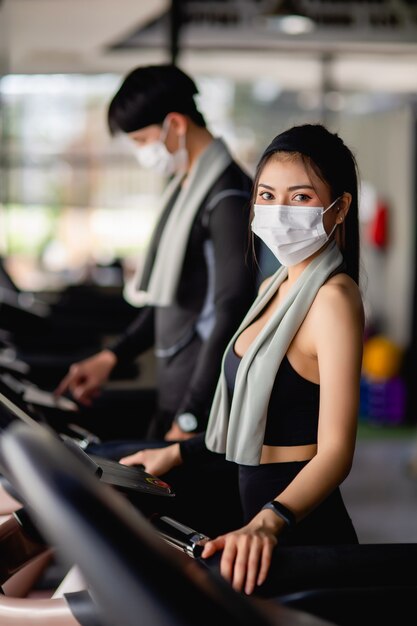 선택적 초점, 스포츠웨어와 스마트 워치 및 흐리게 젊은 남자를 입고 마스크에 젊은 섹시한 여자, 그들은 현대 체육관에서 운동을 위해 디딜 방아에 설정 프로그램을 서있다, 복사 공간