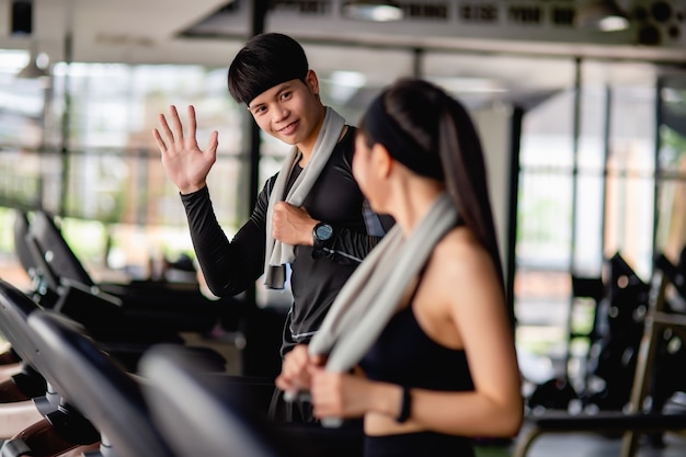 Selective focus, Young sport man smile and raised his hand to greet lovely woman and blurred portrait sexy lady in sportswear on treadmill, they are workout in modern fitness gym, copy space