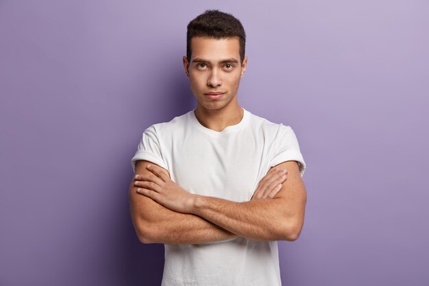 Self assertive serious young guy keeps muscular arms crossed over chest, looks directly, gazes attentively something, dressed in casual white t shirt, looks confident. I am ready listen you