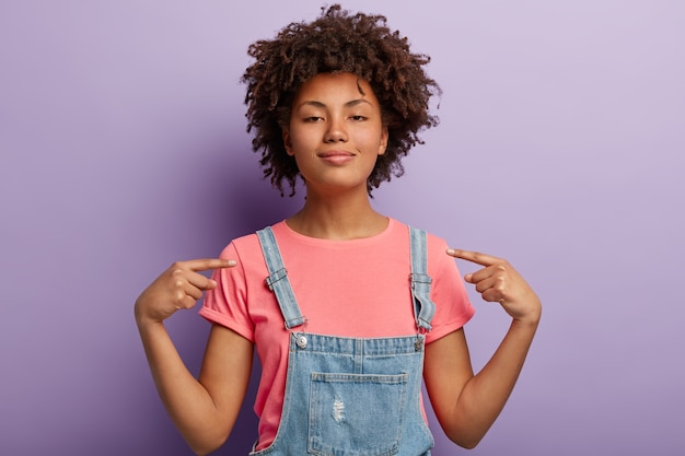Free photo self assured beautiful african american woman feels proud of her deeds, points at herself feels surge of pride, raises head, has dark healthy skin, wears casual clothes, isolated over purple wall