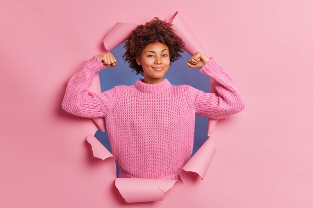 Self assured beautiful young Afro American woman raises arms shows biceps being strong and powerful