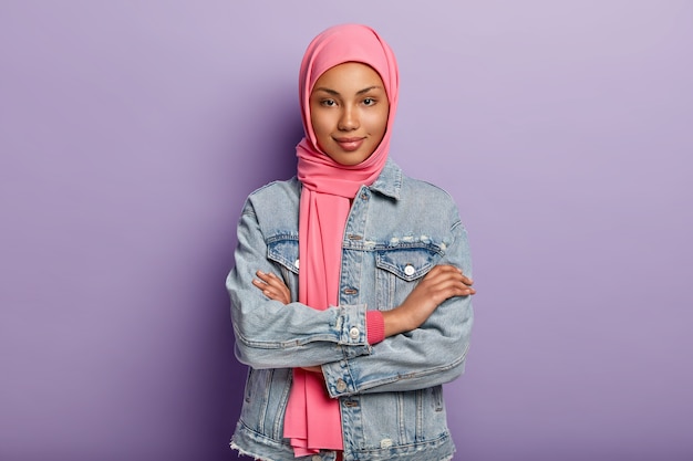 La donna dalla pelle scura sicura di sé ha una visione religiosa musulmana, tiene le braccia incrociate, indossa un velo rosa e un cappotto di jeans, isolata su un muro viola, ascolta le notizie con interesse