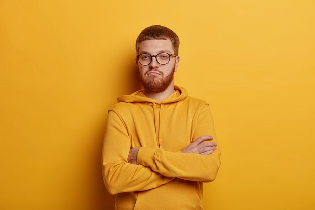 Self assured man with ginger hair and stubble, crosses arms over chest, being confident and boasts about his achievements, dressed casually, poses in yellow, doesnt trust friend looks