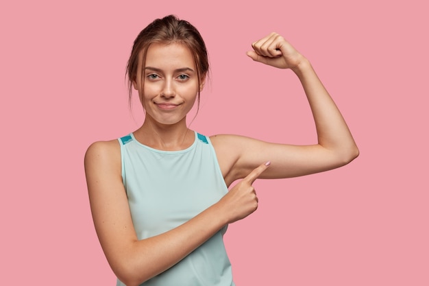 Self assured pretty dark haired sporty woman with pleasant appearance, raises hand and indicates at biceps, confirms women can be also strong, wears light blue vest, isolated over pink wall